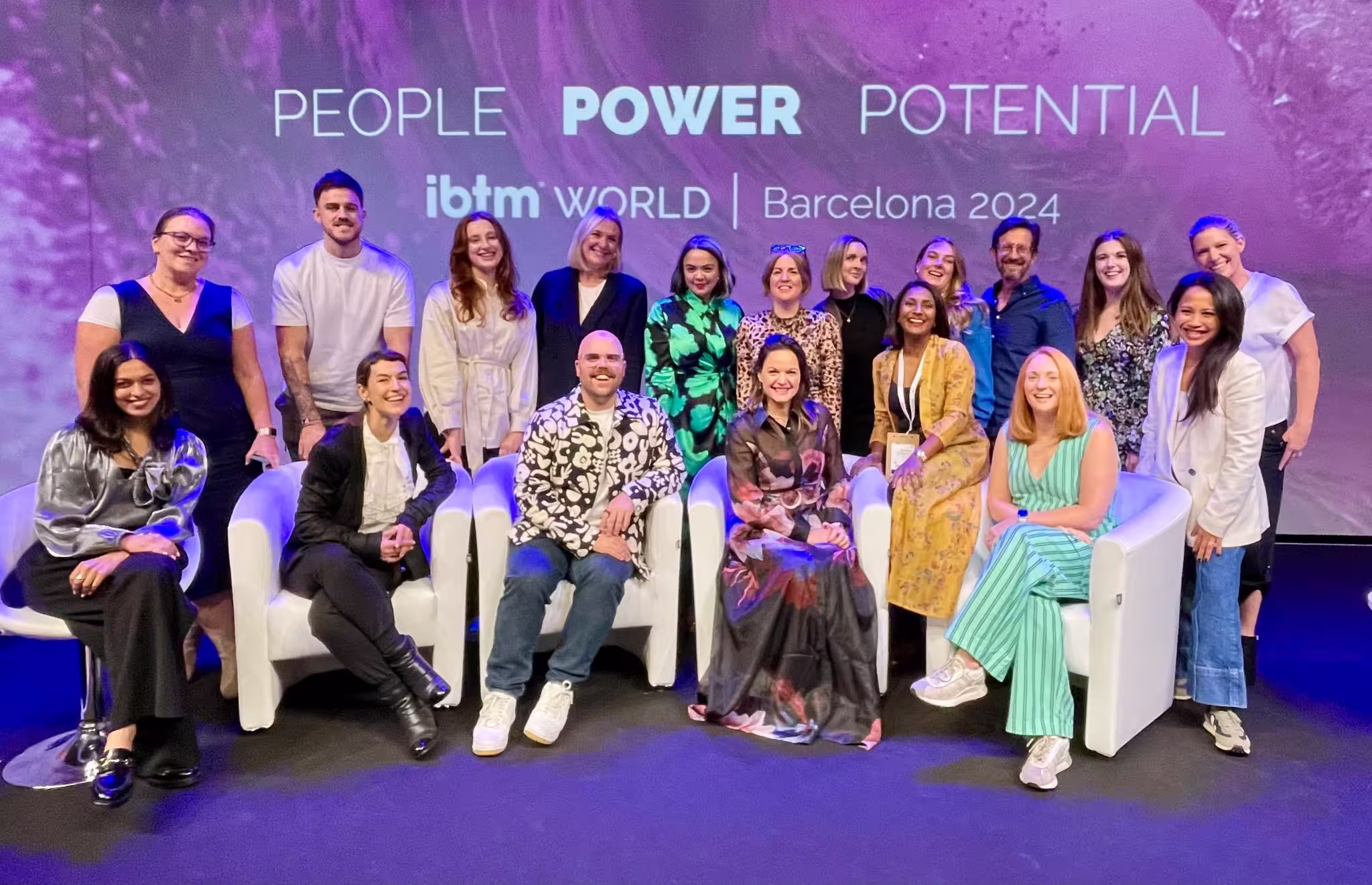 A large group of conference delegates and speakers pose together on stage at IBTM World Barcelona 2024, seated and standing in front of a purple backdrop reading "People Power Potential"