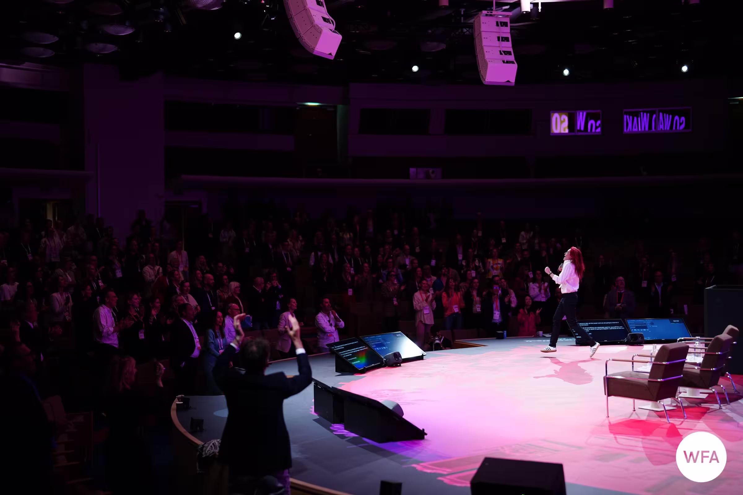 A red-haired speaker commands a large conference stage lit in pink and purple, addressing a packed standing audience at a WFA Global Marketer Week event, as an attendee in the foreground raises their arm enthusiastically.