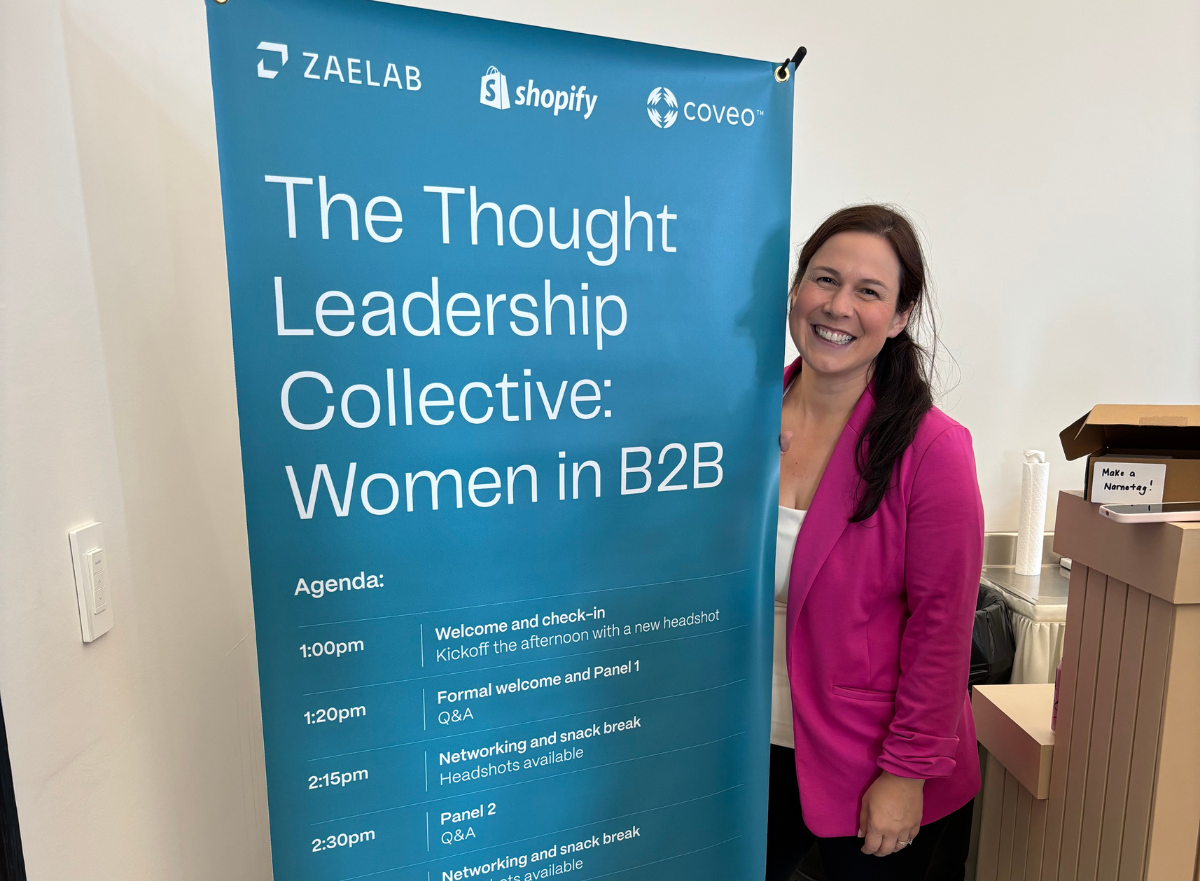 A smiling attendee poses next to the event agenda banner for the Women in B2B Thought Leadership Collective, sponsored by Zaelab, Shopify, and Coveo.