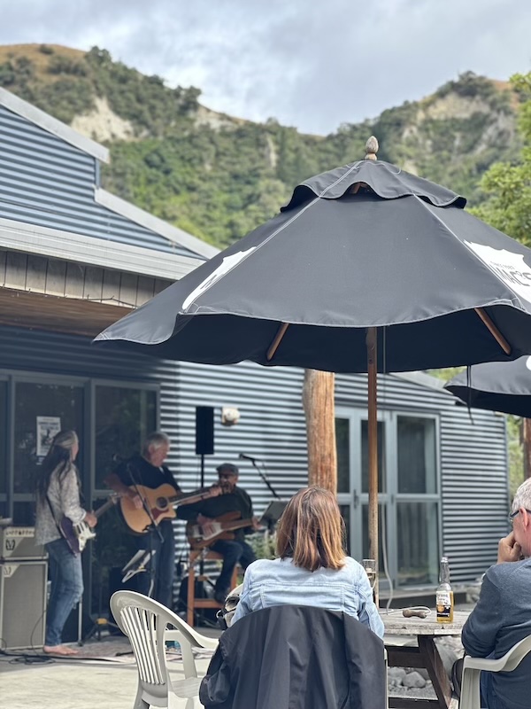 couple sit at a table under umbrella with a beer as they watch a band play in background
