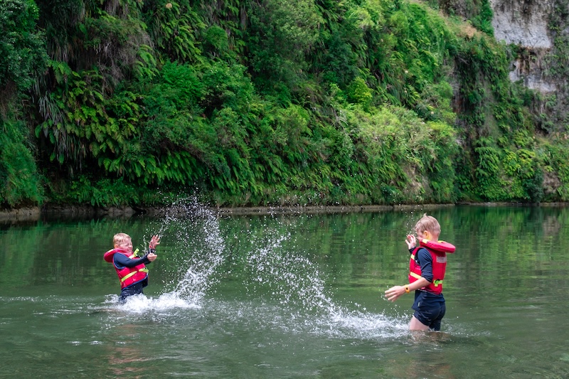 two boys standing splashing eachother in knee deep water on the rangitikei river below steep native lined cliffs
