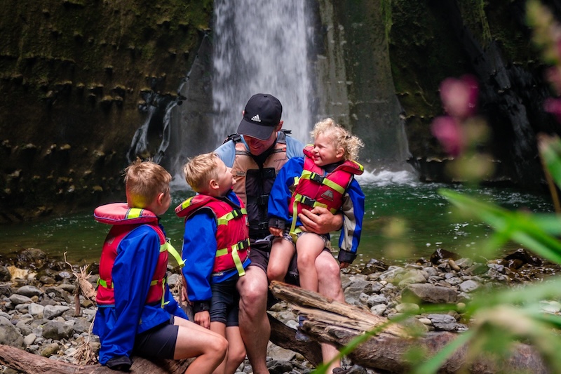 three kids and their dad sit below a small waterfall and pool in a deep narrow gorge on the rangitikei river in new zealand