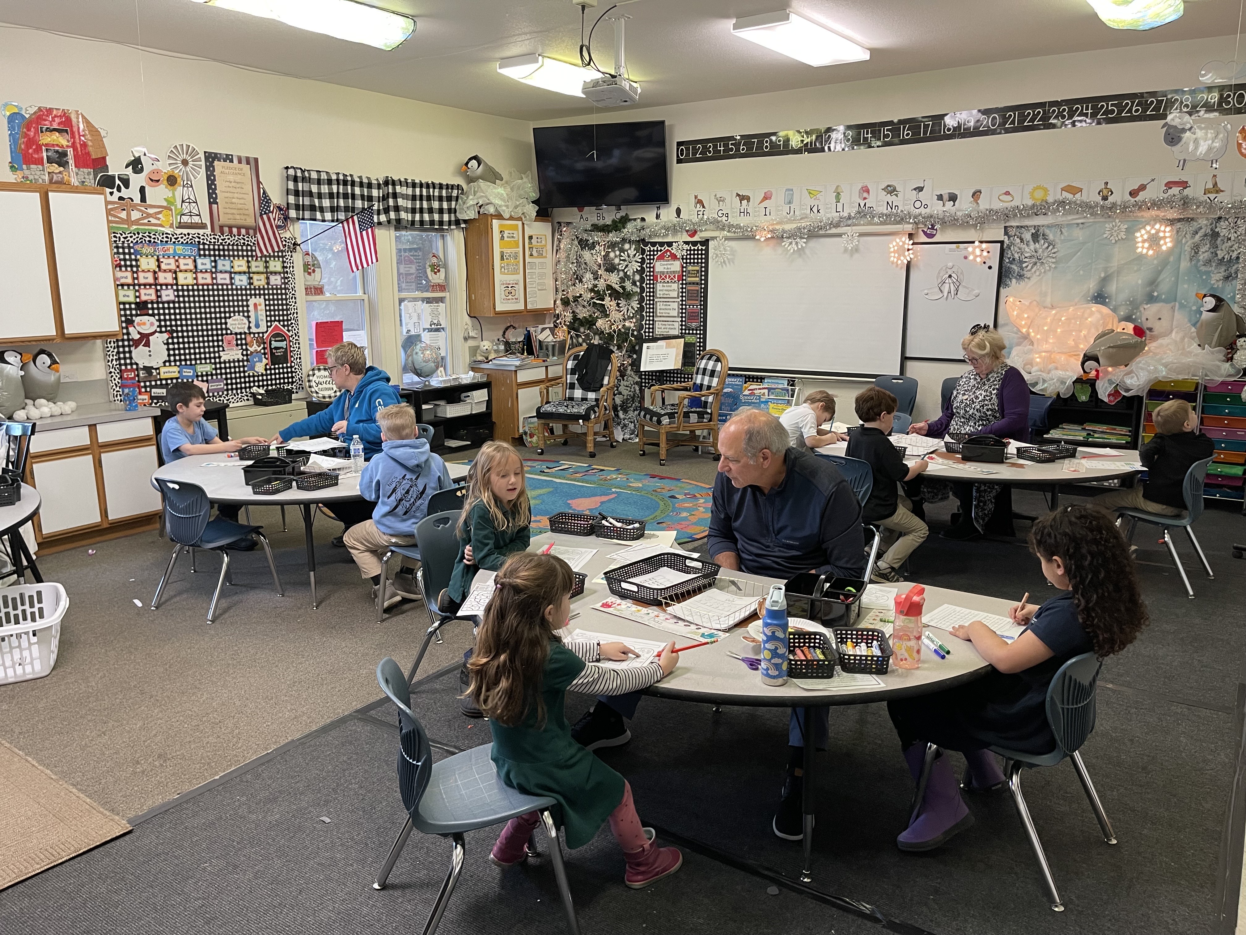 Classroom with children and adults seated at round tables working on activities with educational decor.