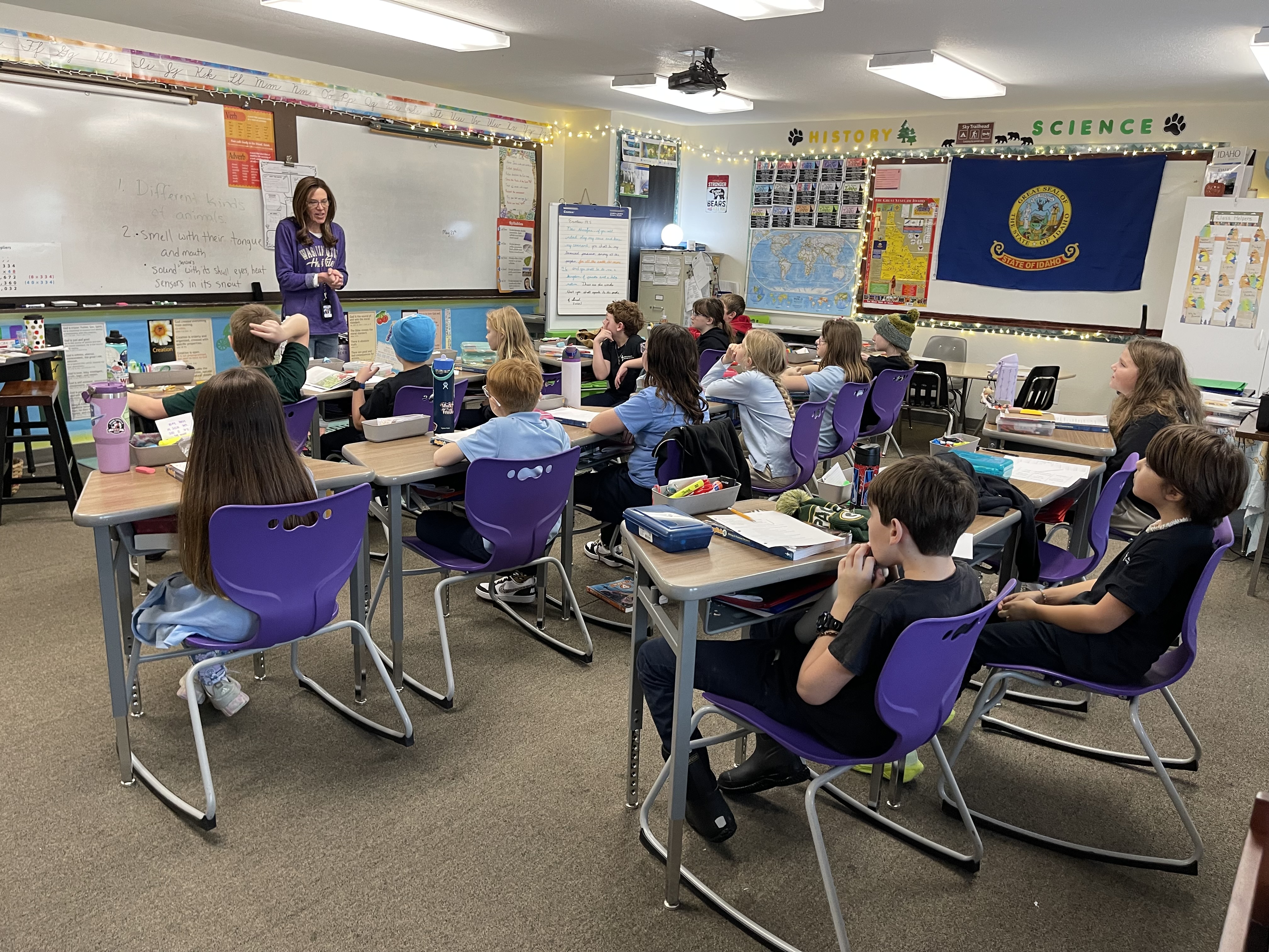 Classroom with a teacher instructing attentive elementary students seated on purple chairs.
