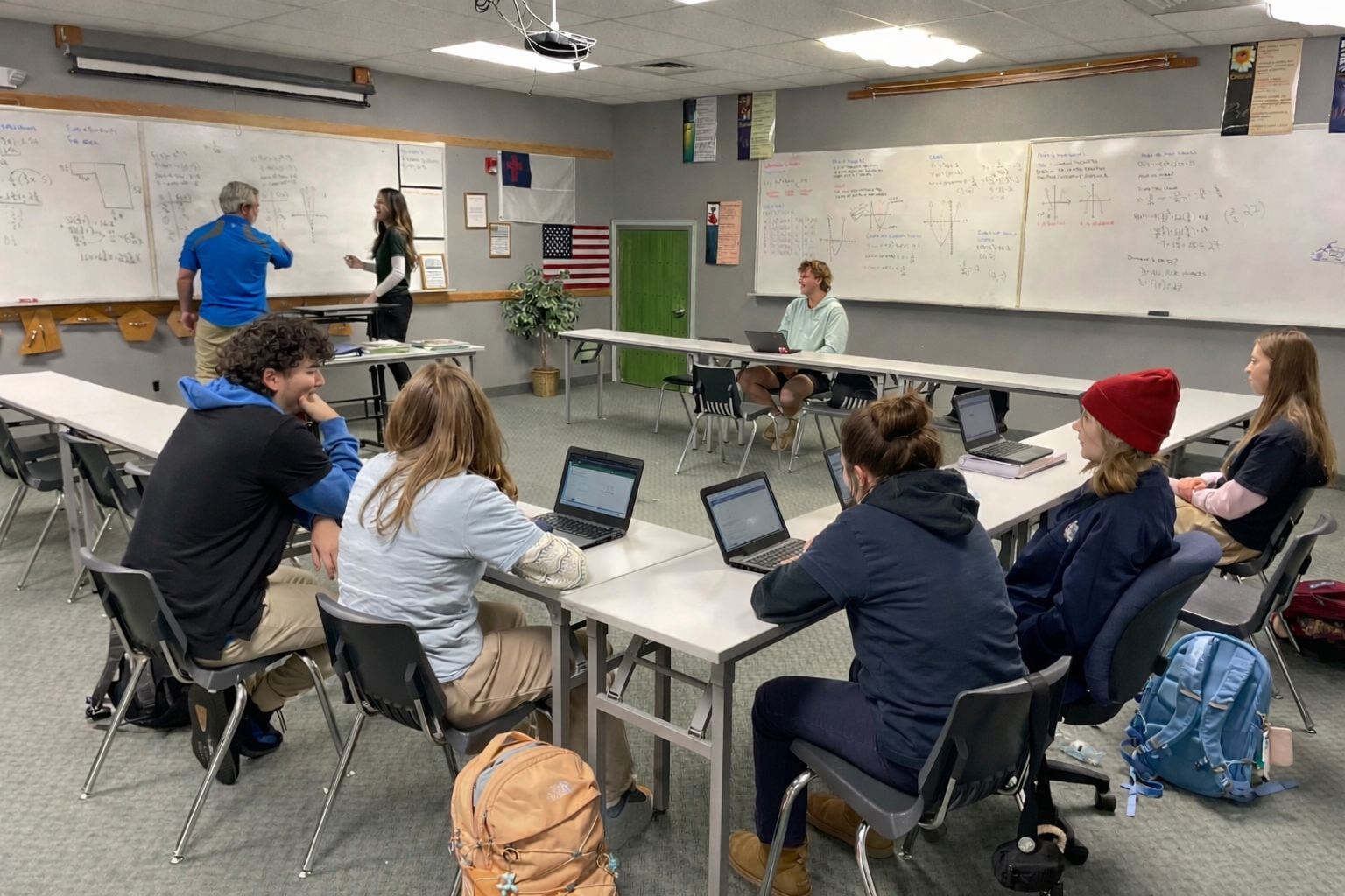 Students seated with laptops in a classroom while teacher discusses math formulas on the whiteboard.