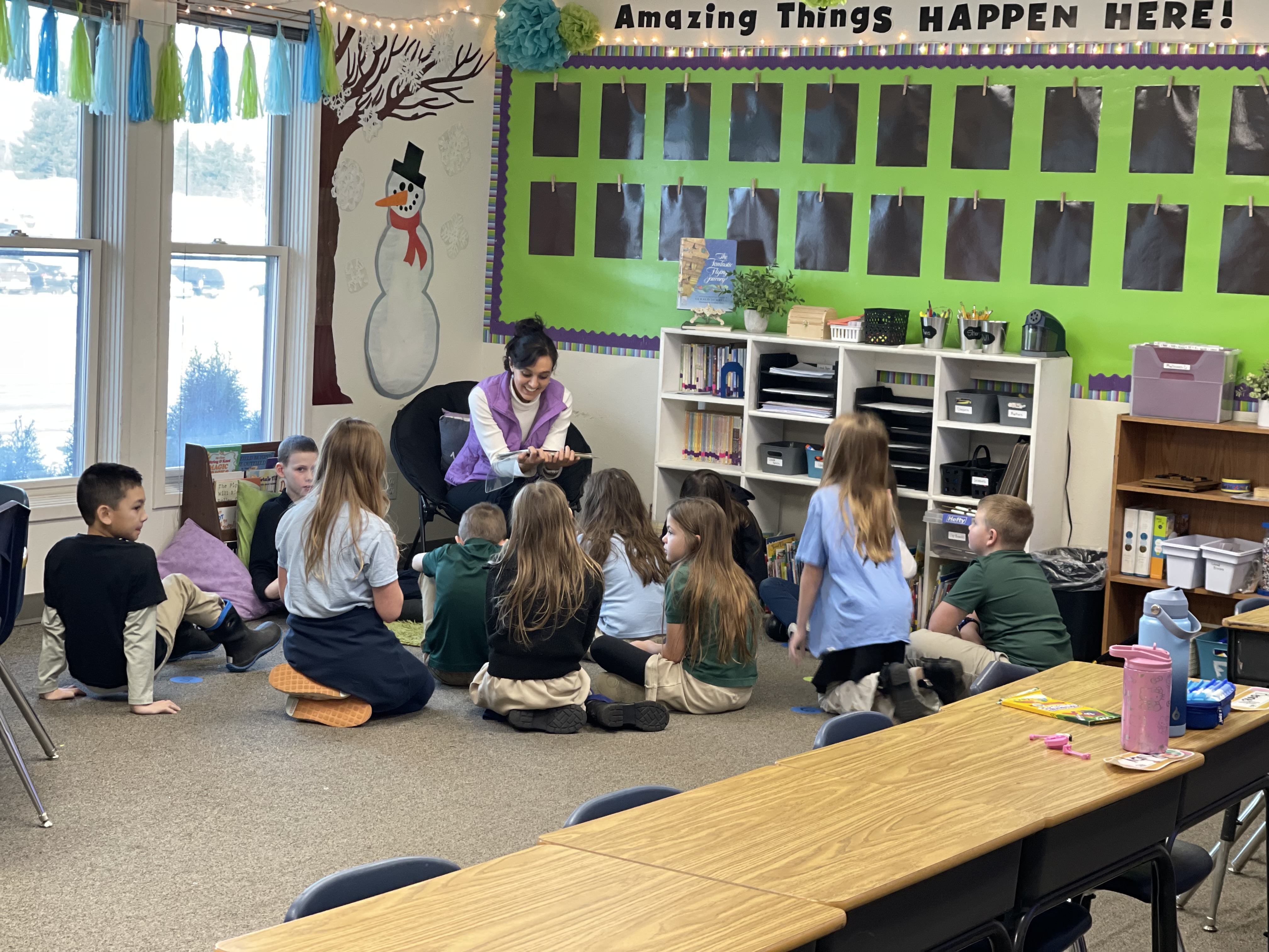 Teacher reading to a group of children sitting on the classroom floor under a sign that says Amazing Things Happen Here.