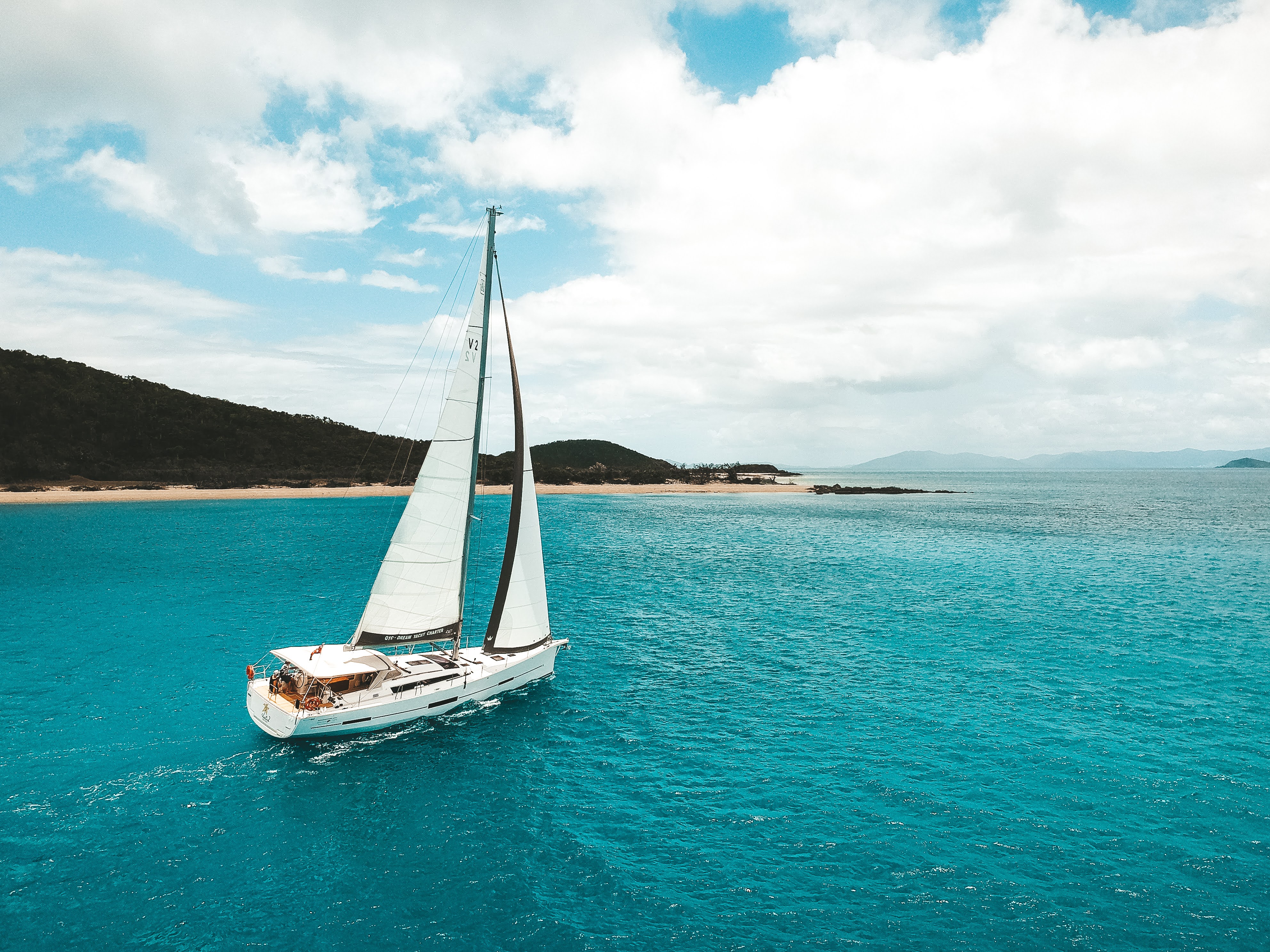 White sailboat cruising on bright turquoise water near a sandy beach with forested hills under a partly cloudy sky.