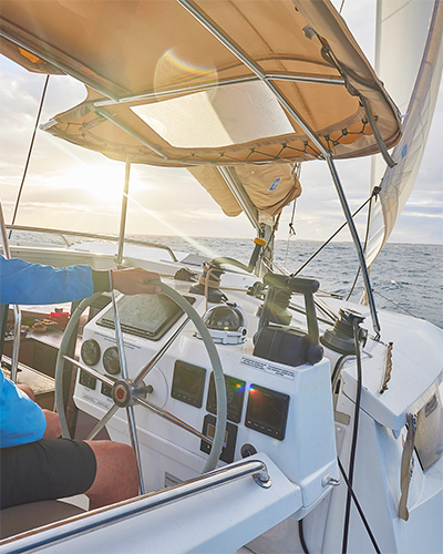 Person steering a sailboat near the helm with calm ocean and sunset in the background.