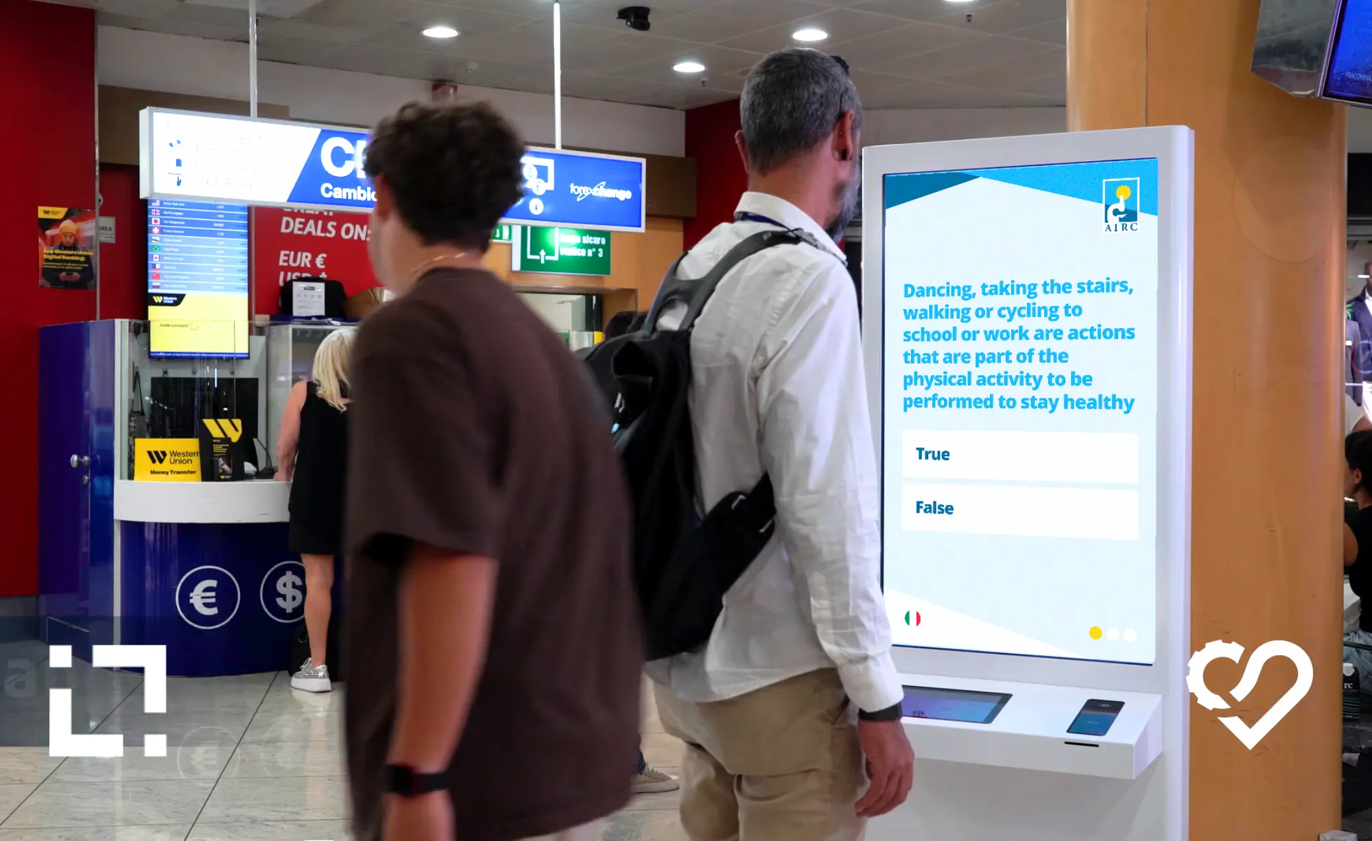 Viaggiatori davanti al totem interattivo AIRC con quiz sulla prevenzione del cancro