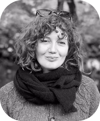 Smiling woman with curly hair, glasses on head, wearing a scarf and sweater outdoors.