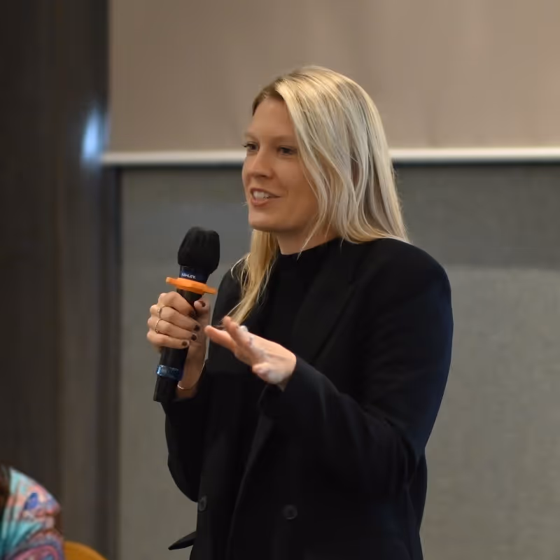 Blonde woman in black jacket speaking into a microphone while gesturing with her hand.