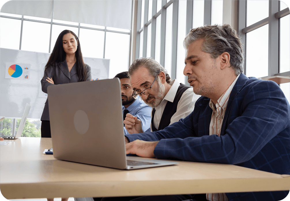 Four colleagues in a meeting room focused on a laptop, with charts on a whiteboard behind them.