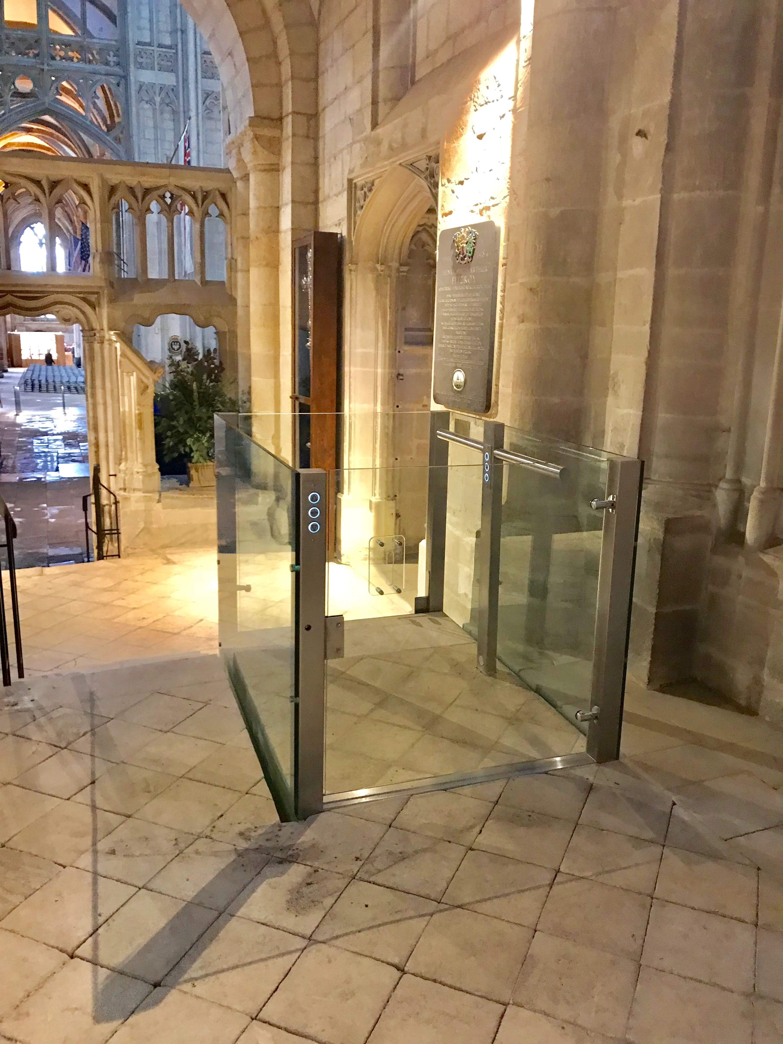 Bespoke DDA access step lift providing wheelchair access between levels in a public building. The step lift has steel painted framework with glass side panels and gate