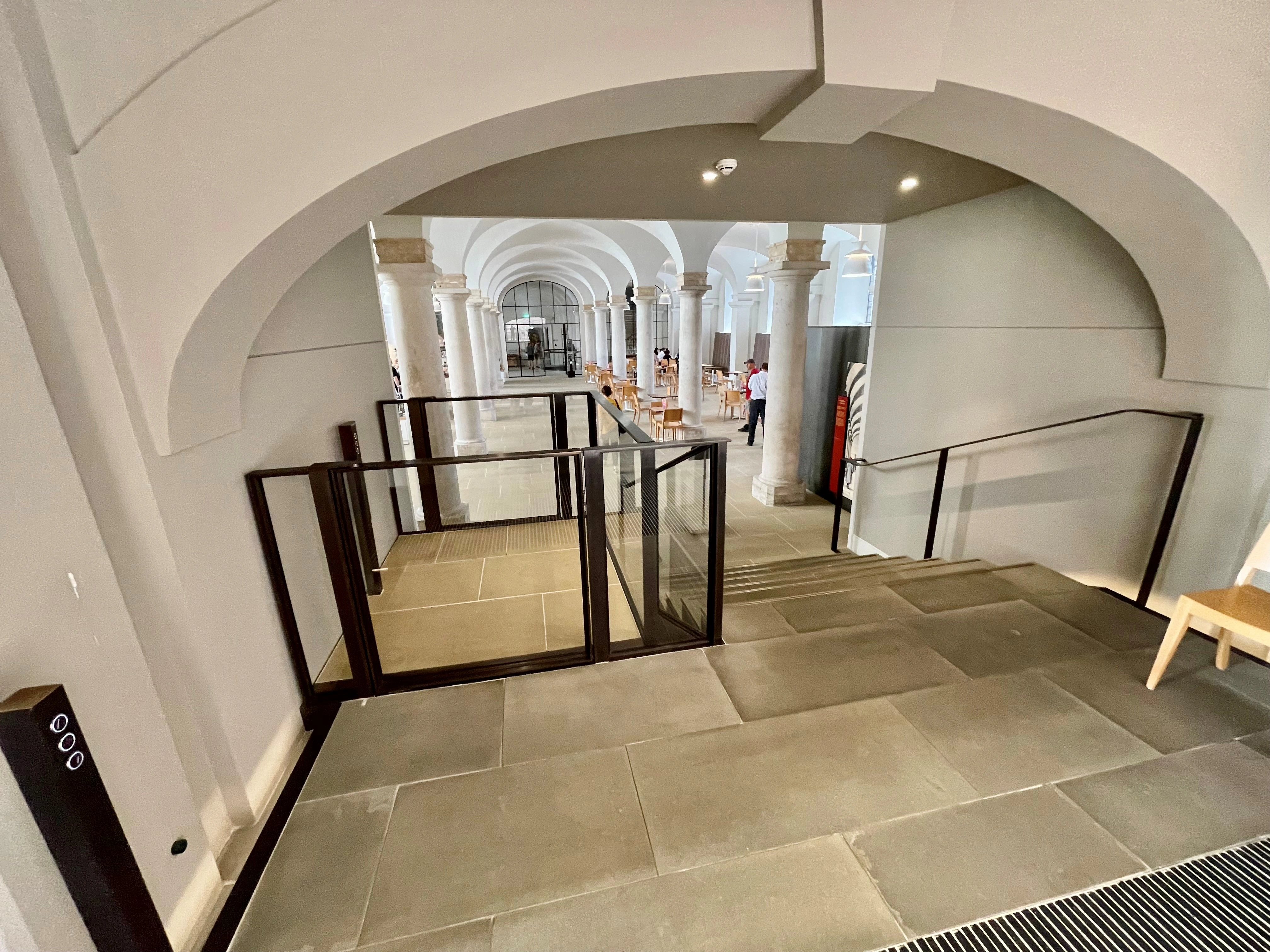 Bespoke DDA access step lift providing wheelchair access between levels in a public building. The step lift has steel painted framework with glass side panels and gate