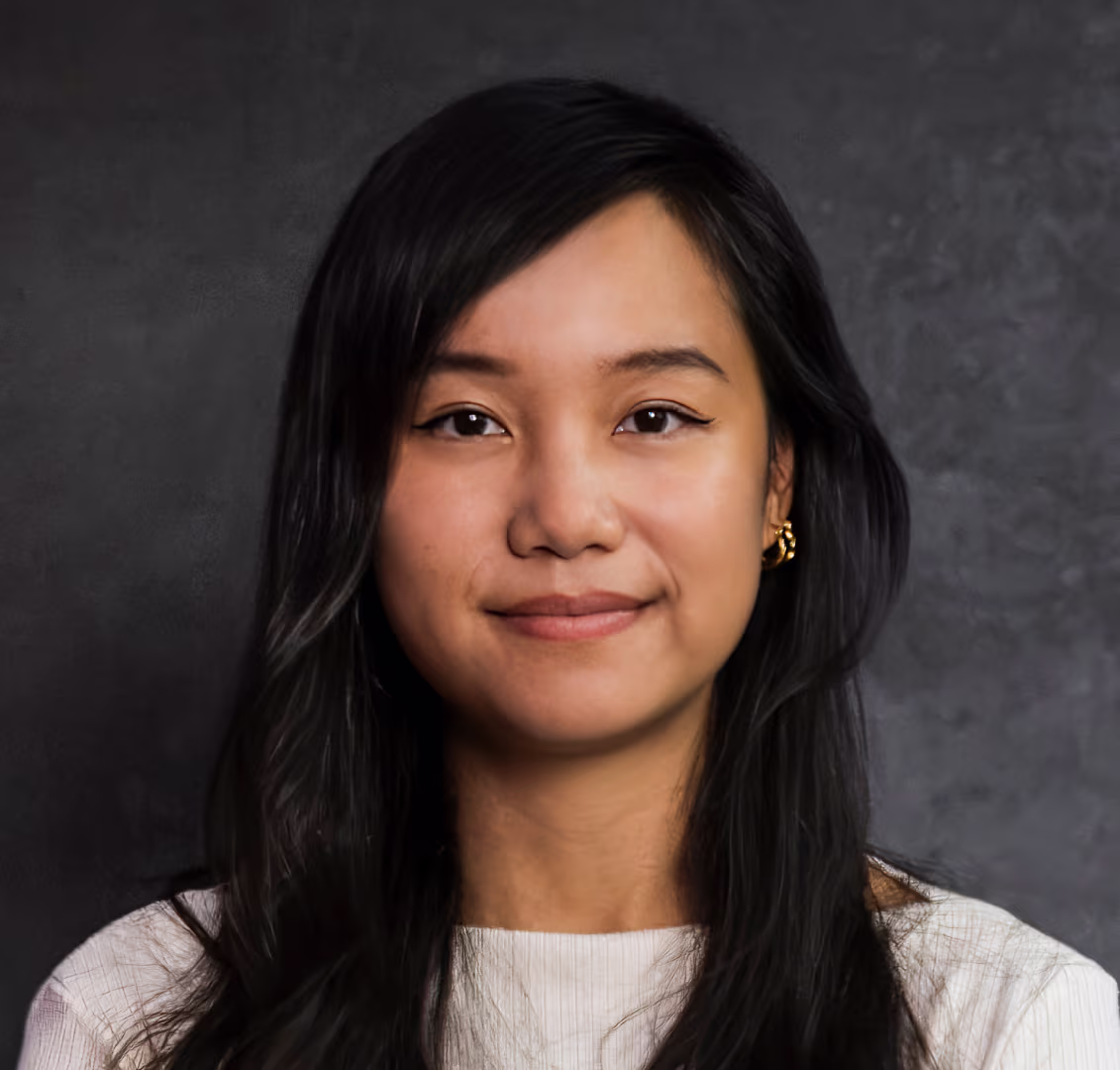 Portrait einer Frau mit langen schwarzen Haaren, die ein helles Oberteil vor dunklem Hintergrund trägt und leicht lächelt.