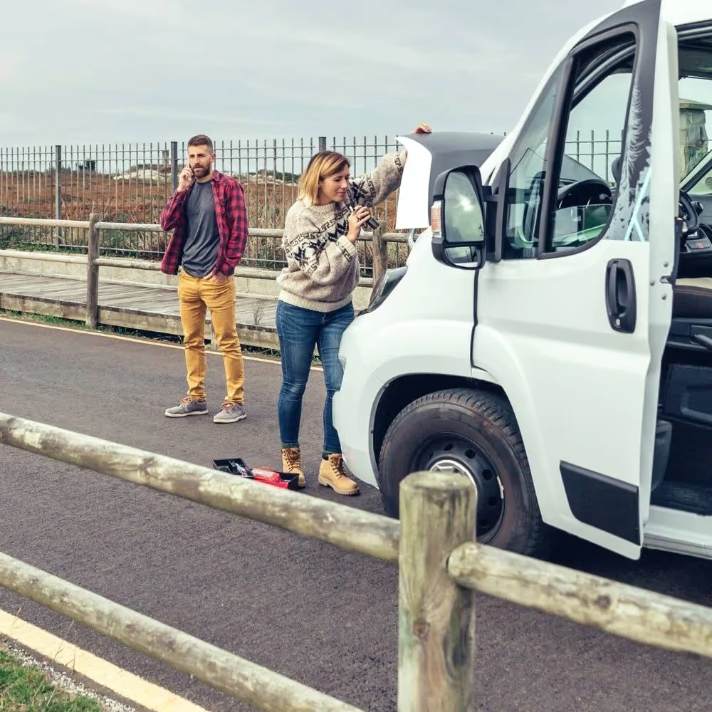 A woman checks under the open hood of a van while a man watches nearby.