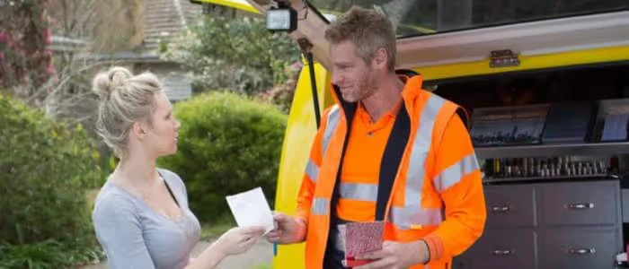 A woman hands a piece of paper to a worker in an orange safety vest near a van.