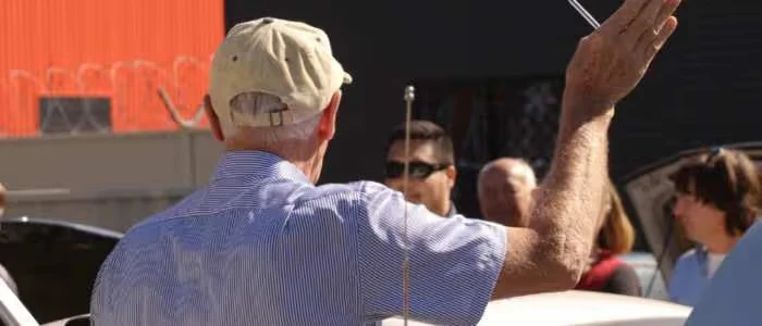 An older man wearing a cap gestures with his hand while talking to a small group.