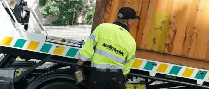 A worker in a high-visibility jacket operates a truck loading a large orange container.