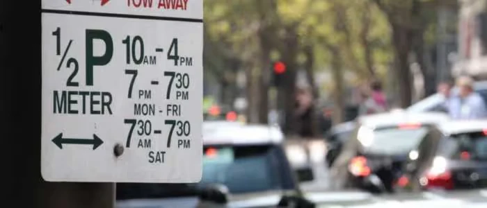 Parking meter sign showing hours, with busy street and blurred cars in the background.