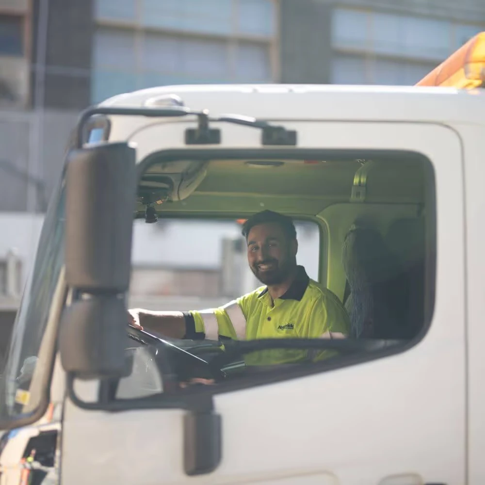 A person in a high-visibility shirt is smiling while sitting in a truck driver's seat.