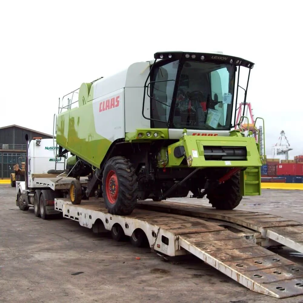 Green and white CLAAS agricultural harvester loaded on a flatbed trailer truck at an industrial port area.