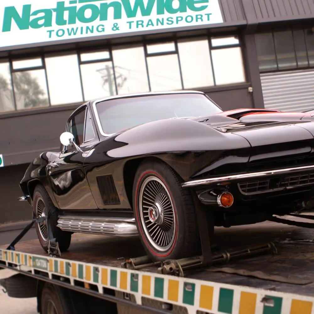 A classic black sports car is loaded onto a tow truck in front of a transport company.