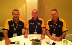 Three men in matching polo shirts sit at a table with water glasses and a jug.