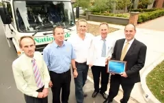 Five men in formal attire standing outdoors near a truck, one holding a framed certificate.