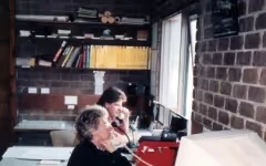 Two women sitting and talking on telephones in a brick-walled office with shelves and a large window.
