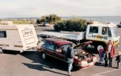 A roadside assistance truck helps a car with an open hood, towing a small camper.