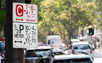 Street parking sign shows meter times and tow-away hours, with cars and trees in the background.