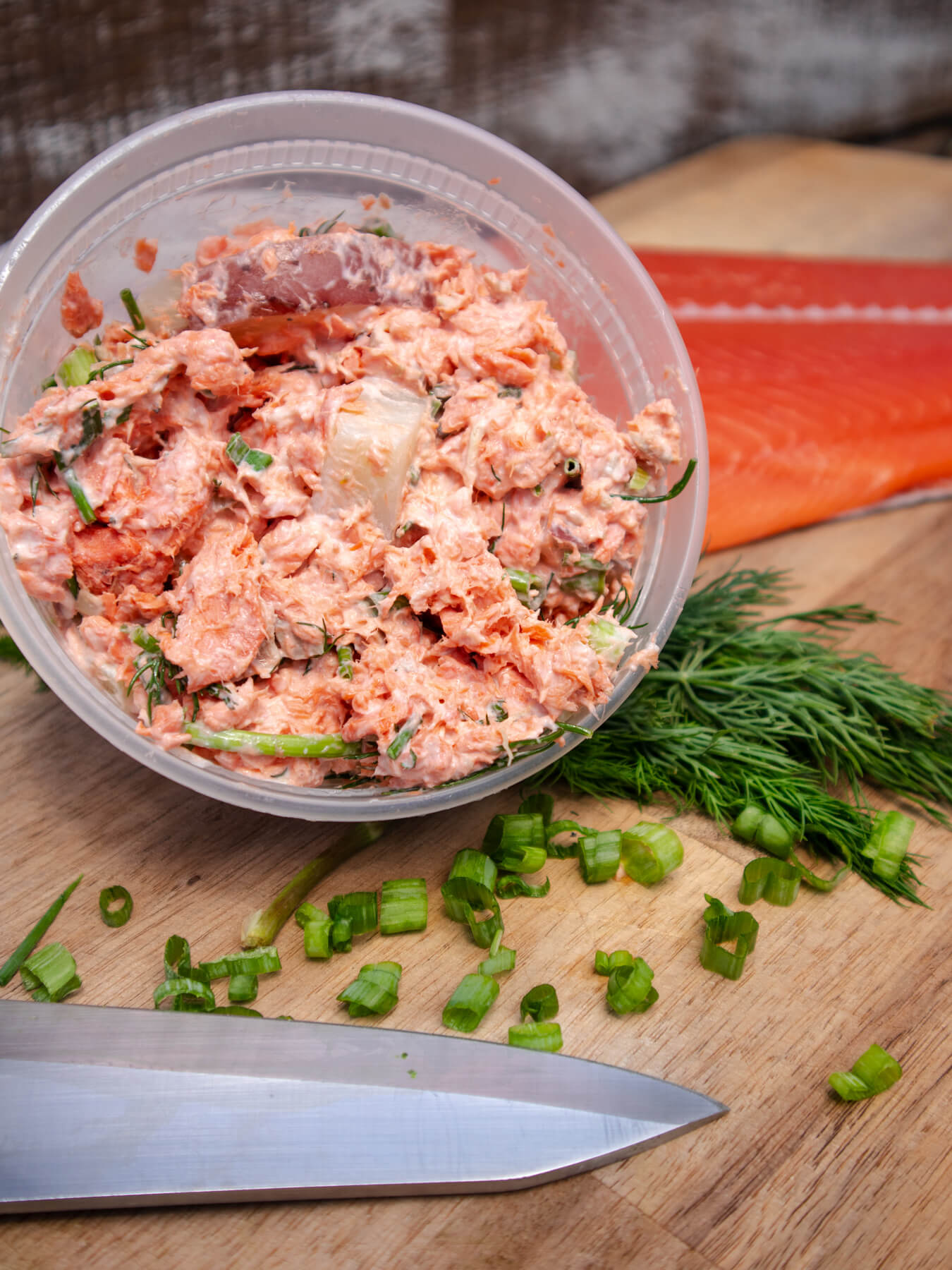 Close up of Salmon Potato Salad in container with fresh green onions and dill next to it with a Salmon filet in the background and knife in the foreground