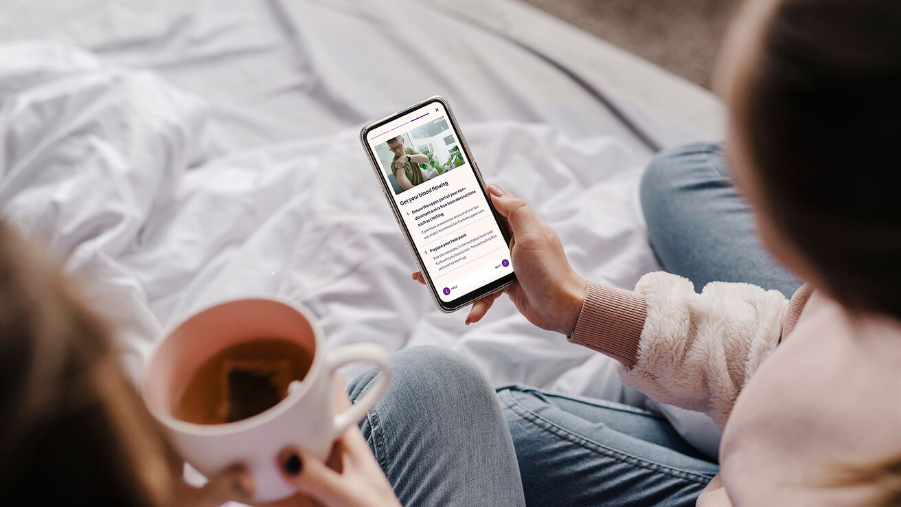 A lady is looking at a phone in bed and reading the instructions for taking a blood sample. Another lady is sitting next to her holding a cup of tea