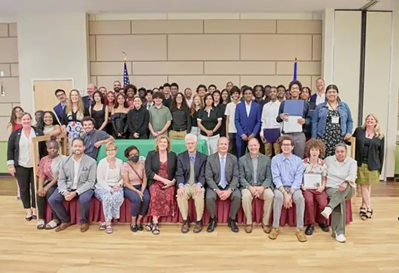 A large diverse group of men and women gathered for a formal recognition or graduation event, posing indoors with flags in the background.