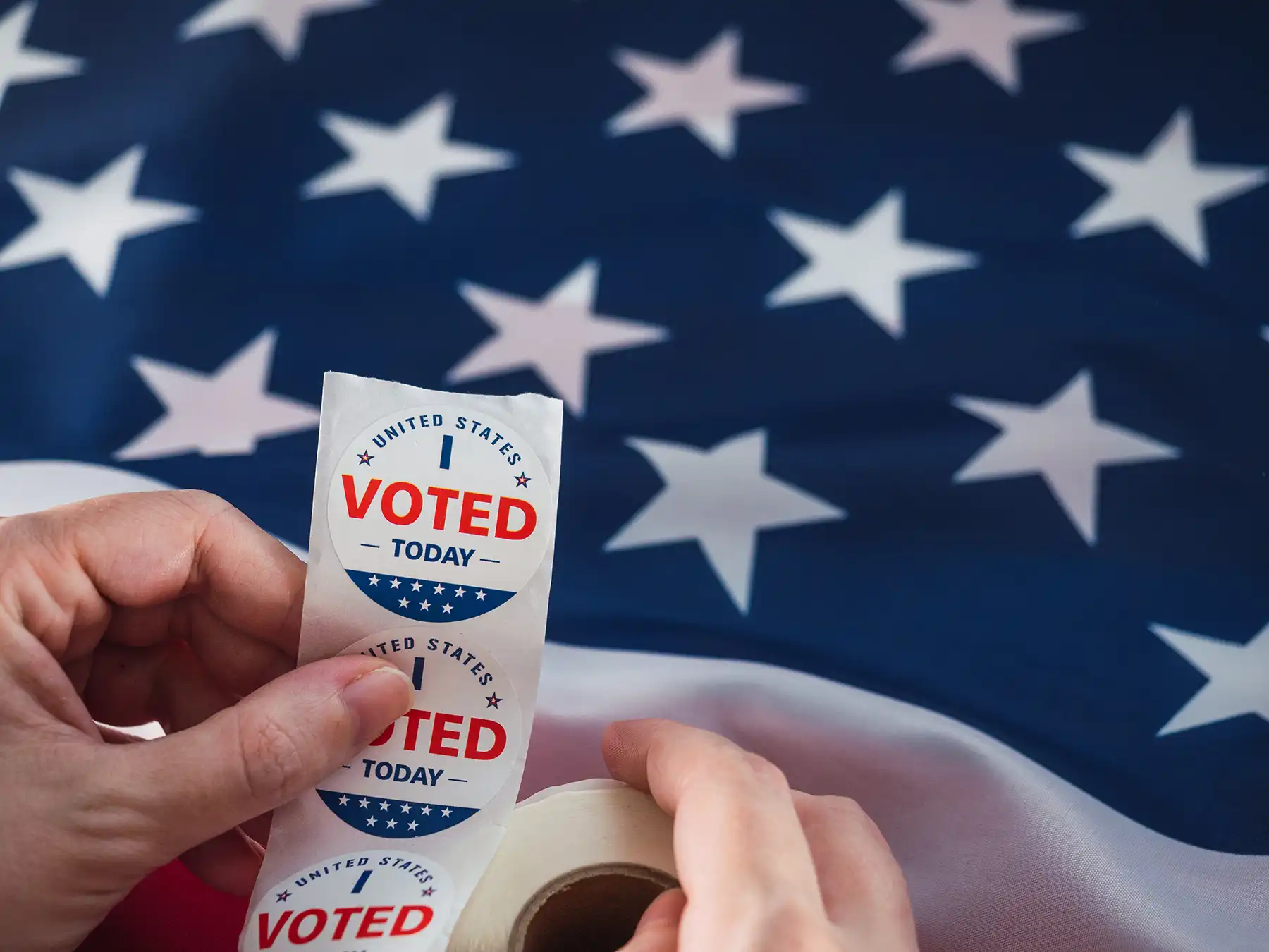 A person holds a roll of "I Voted Today" stickers with an American flag in the background, symbolizing voter participation in United States elections.