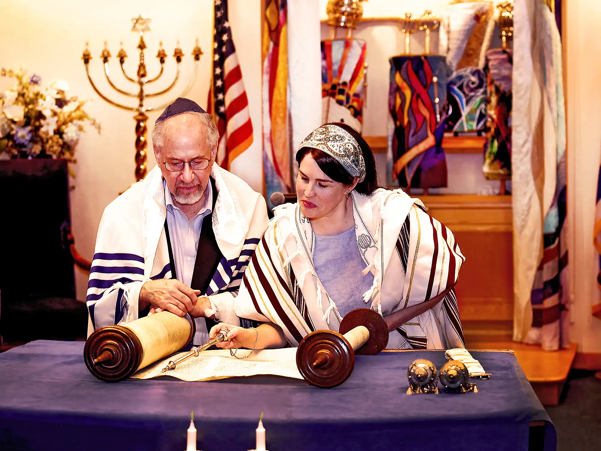 Two rabbis read from the Torah during a service, with a menorah and American flag visible in the background, a symbol of Jewish faith.