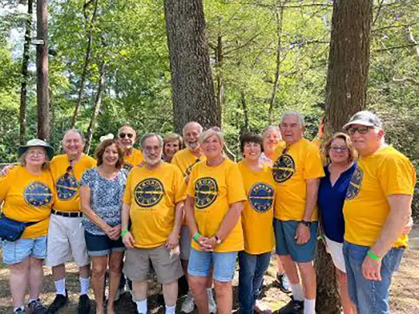 A group of people in yellow event t-shirts pose together outdoors in a wooded area, enjoying a community gathering on a sunny day.