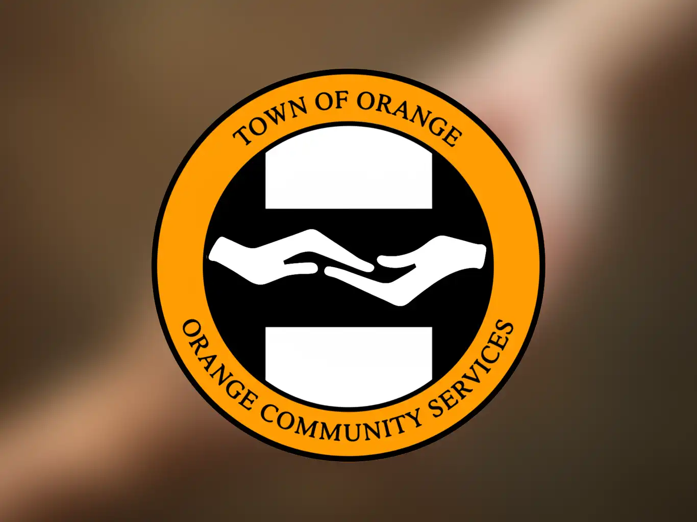 Logo for Town of Orange Community Services, featuring white hands in a black circle with an orange border, on a blurred background.