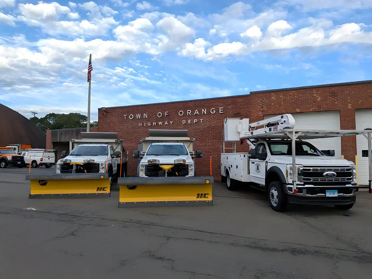 Three new Town of Orange Highway Department vehicles with plows and a bucket truck parked in front of department building.