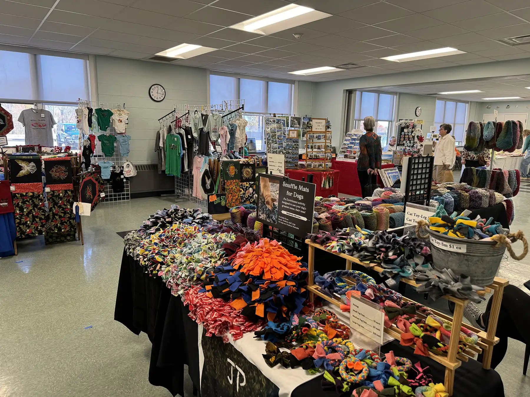 Local vendors display handmade crafts, colorful textiles, and pet snuffle mats at an indoor community craft fair with shoppers browsing tables.
