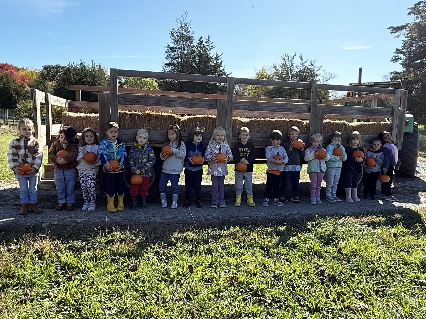 A preschool class of children, about 15 of them, stands in a line outdoors on a sunny day, holding small pumpkins in front of a hayride wagon.