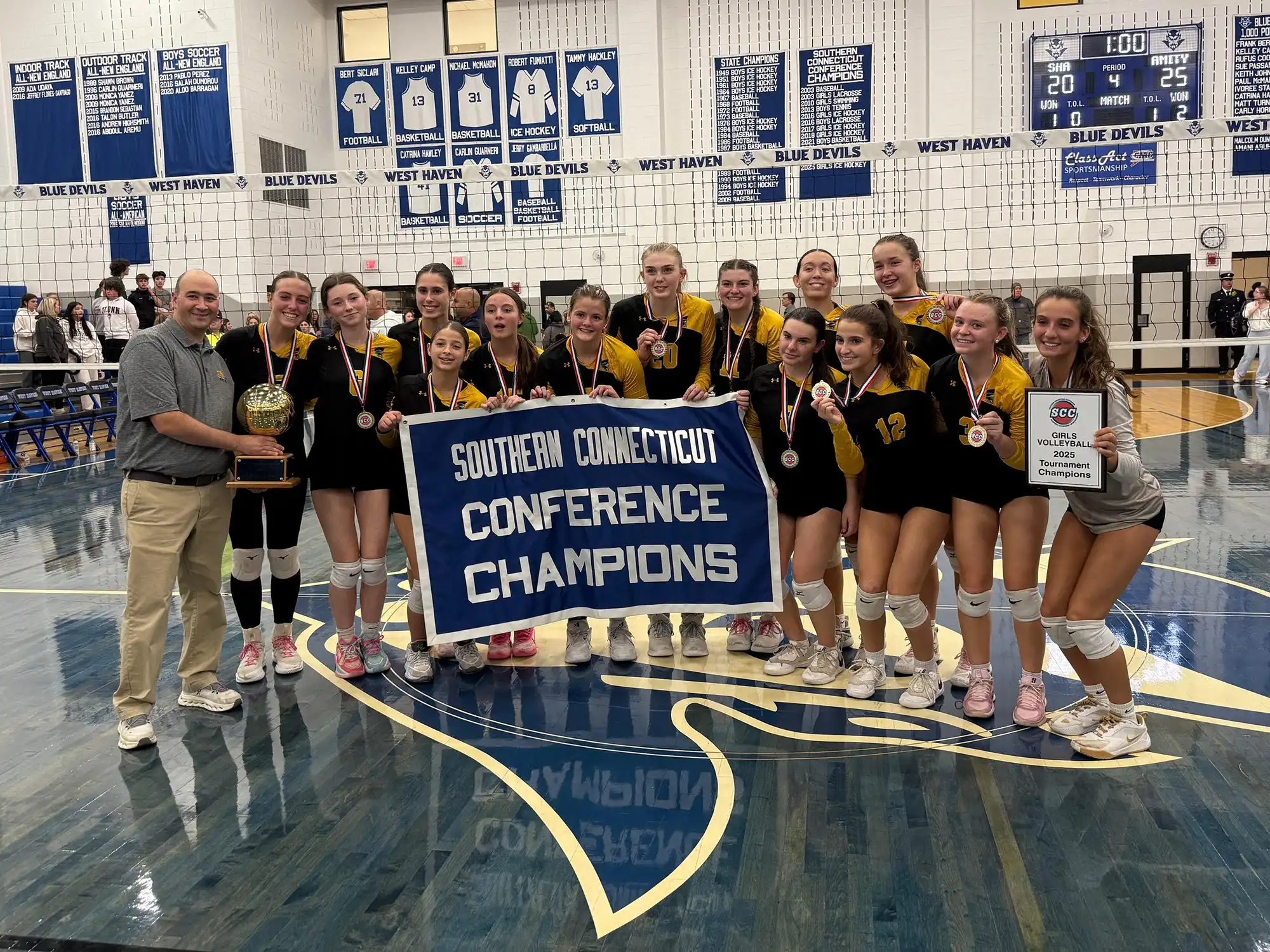 Amity girls volleyball team and coach celebrate winning the SCC championship, holding a trophy, banner, medals, and a plaque on the court.