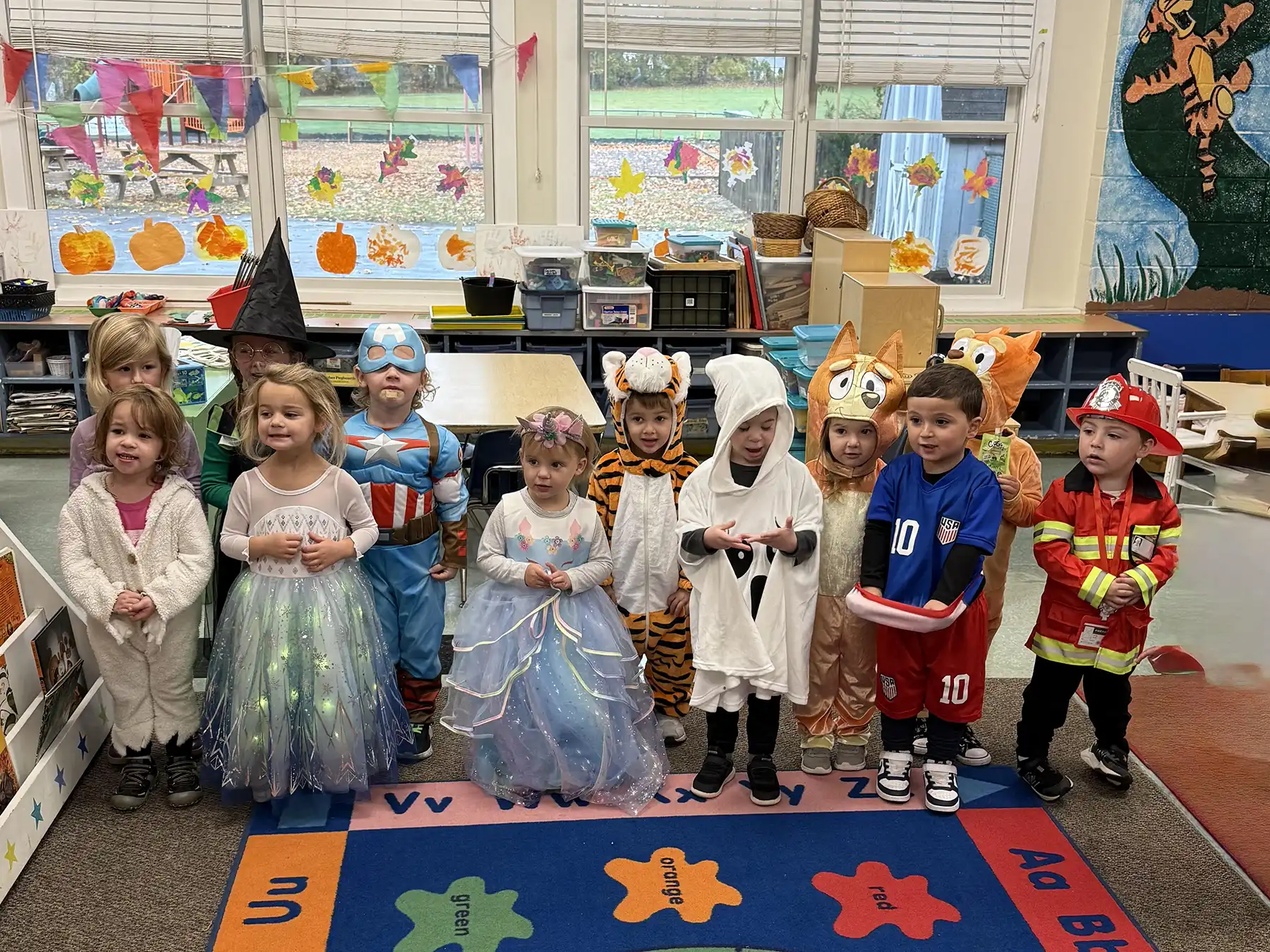 A group of OCNS preschool children in costumes pose for a Halloween parade in a decorated classroom with festive artwork and colorful fall decorations.