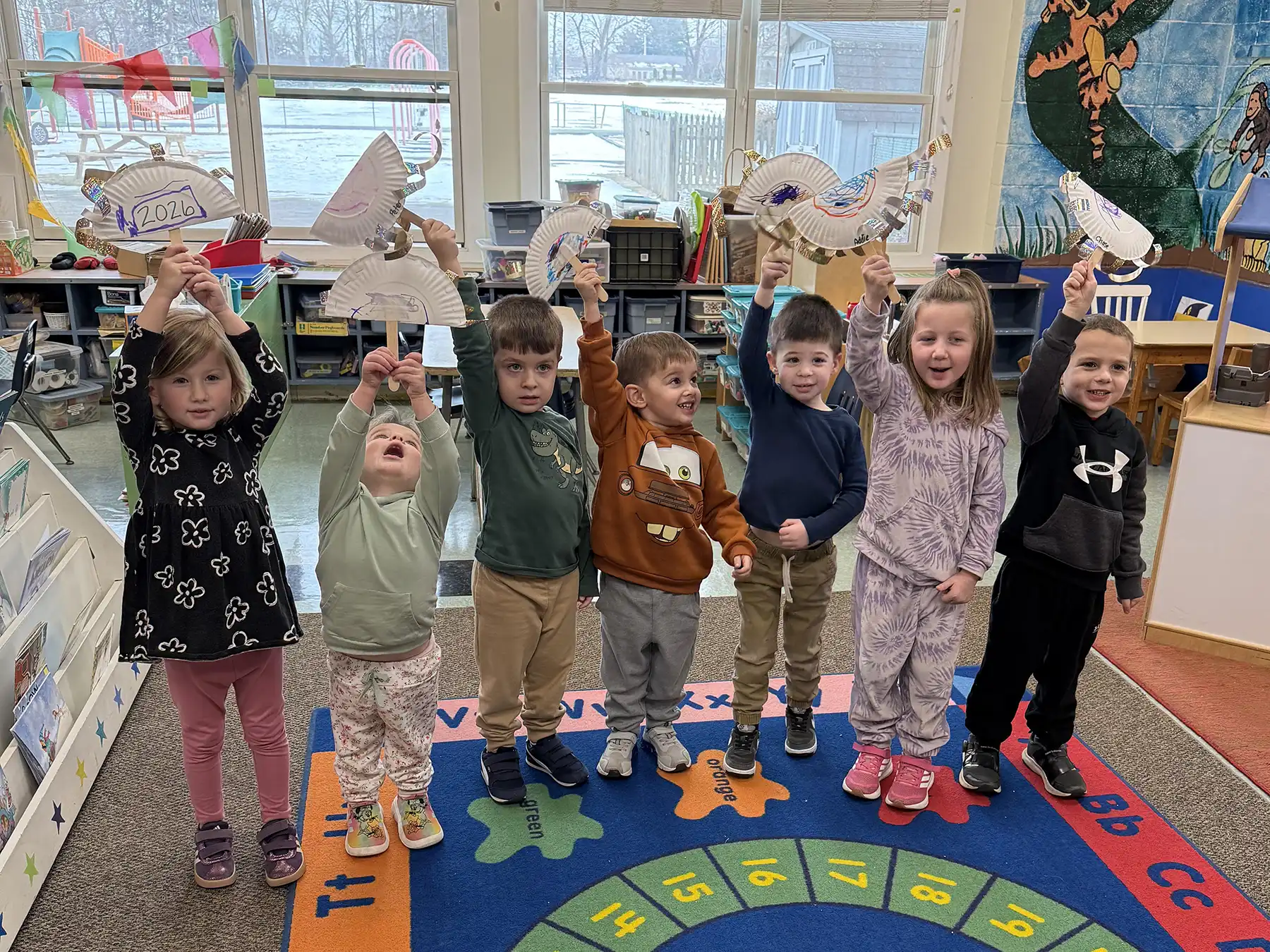 Seven OCNS preschoolers holding paper plate crafts that say 2026 to celebrate the New Year. The children are standing on a colorful rug in a classroom.