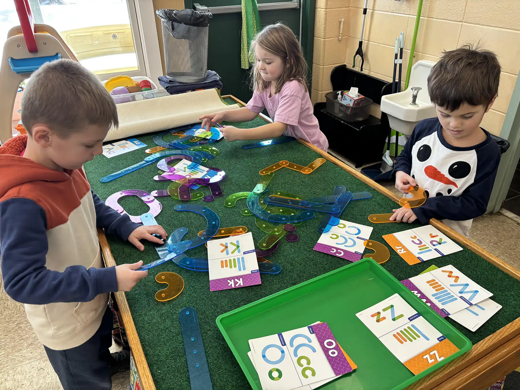 Three children at Orange Community Nursery School use colorful shapes to build letters, practicing literacy in a 5-day kindergarten readiness program.