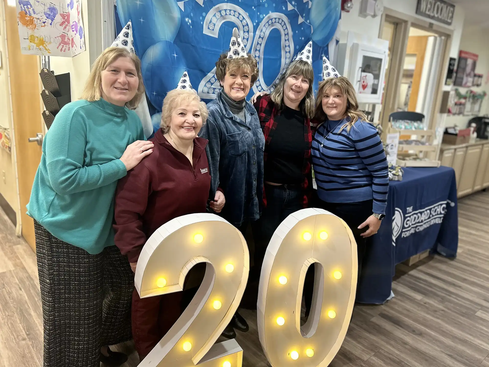 Five staff members wearing party hats pose behind large light-up "20" numbers at a Goddard School anniversary event featuring blue balloons and decorations.