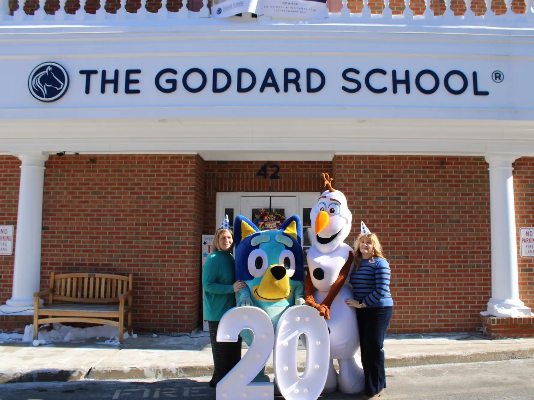 Two staff members pose with Bluey and Olaf costumed characters in front of The Goddard School building, celebrating a 20th anniversary with light-up signs.