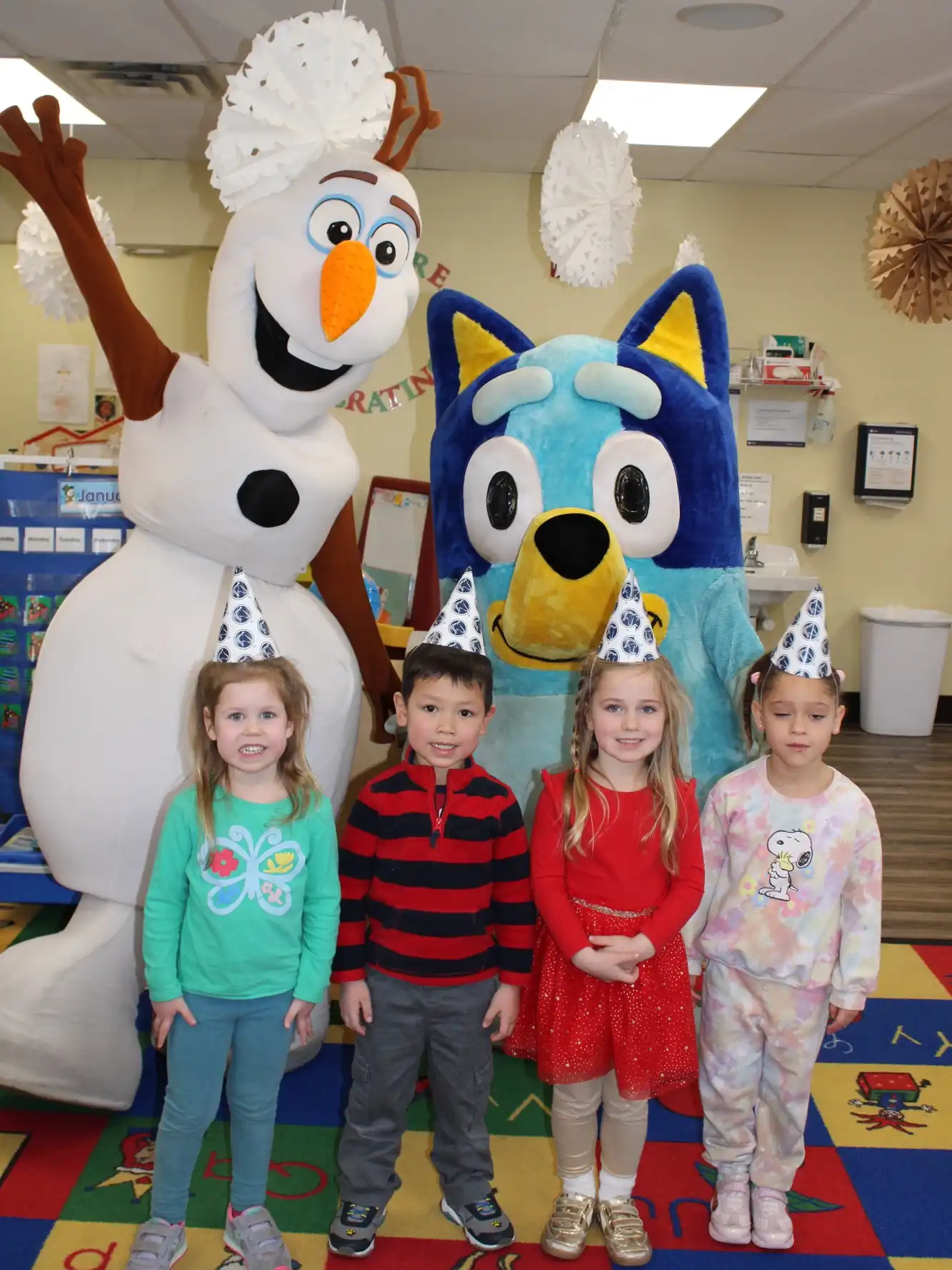 Four children in party hats stand with Bluey and Olaf costumed characters inside a Goddard School classroom during a 20th-anniversary celebration.
