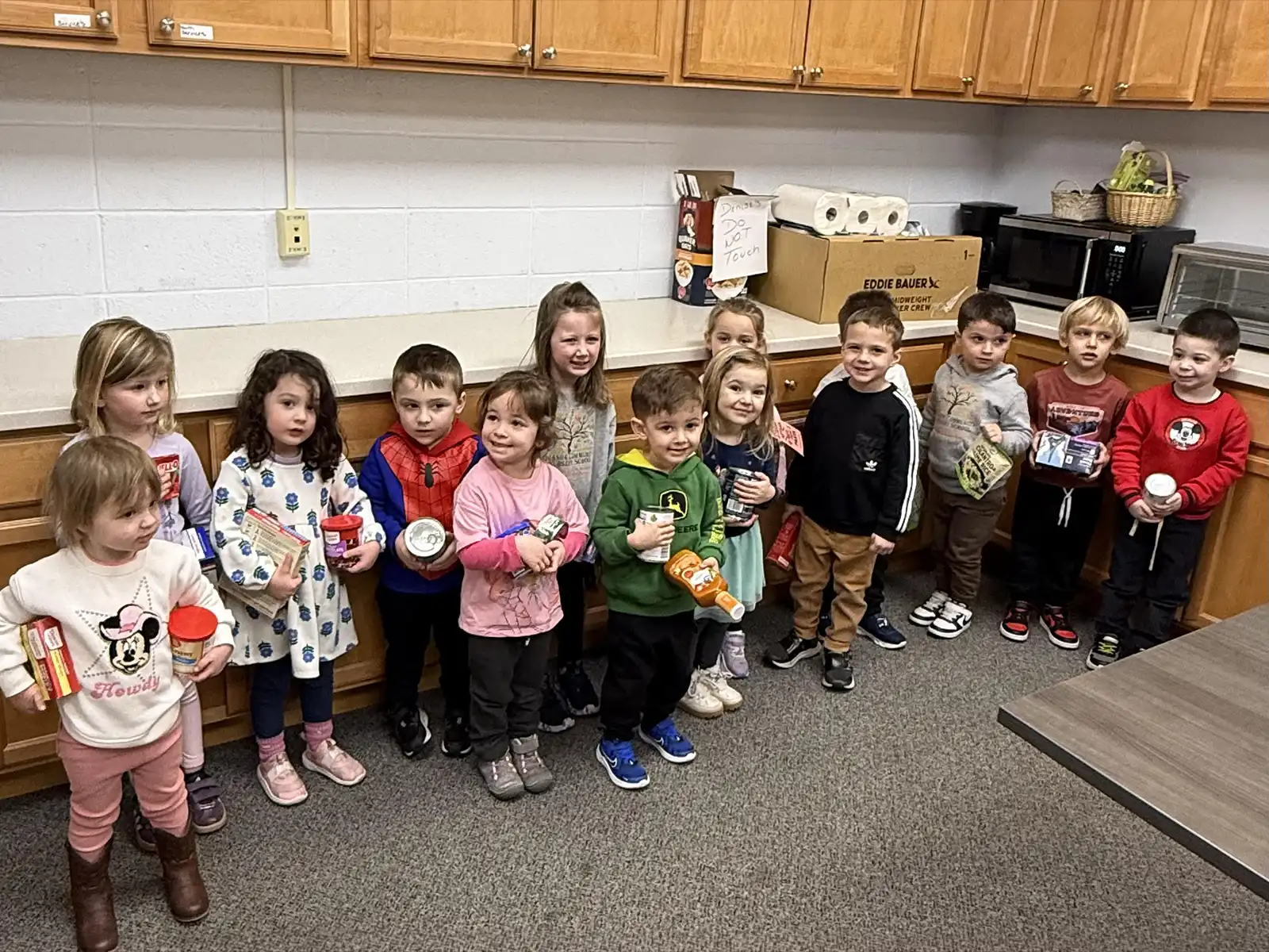 Orange Community Nursery School students stand in a kitchen holding canned goods and boxes of food for the Friendship Food Drive.